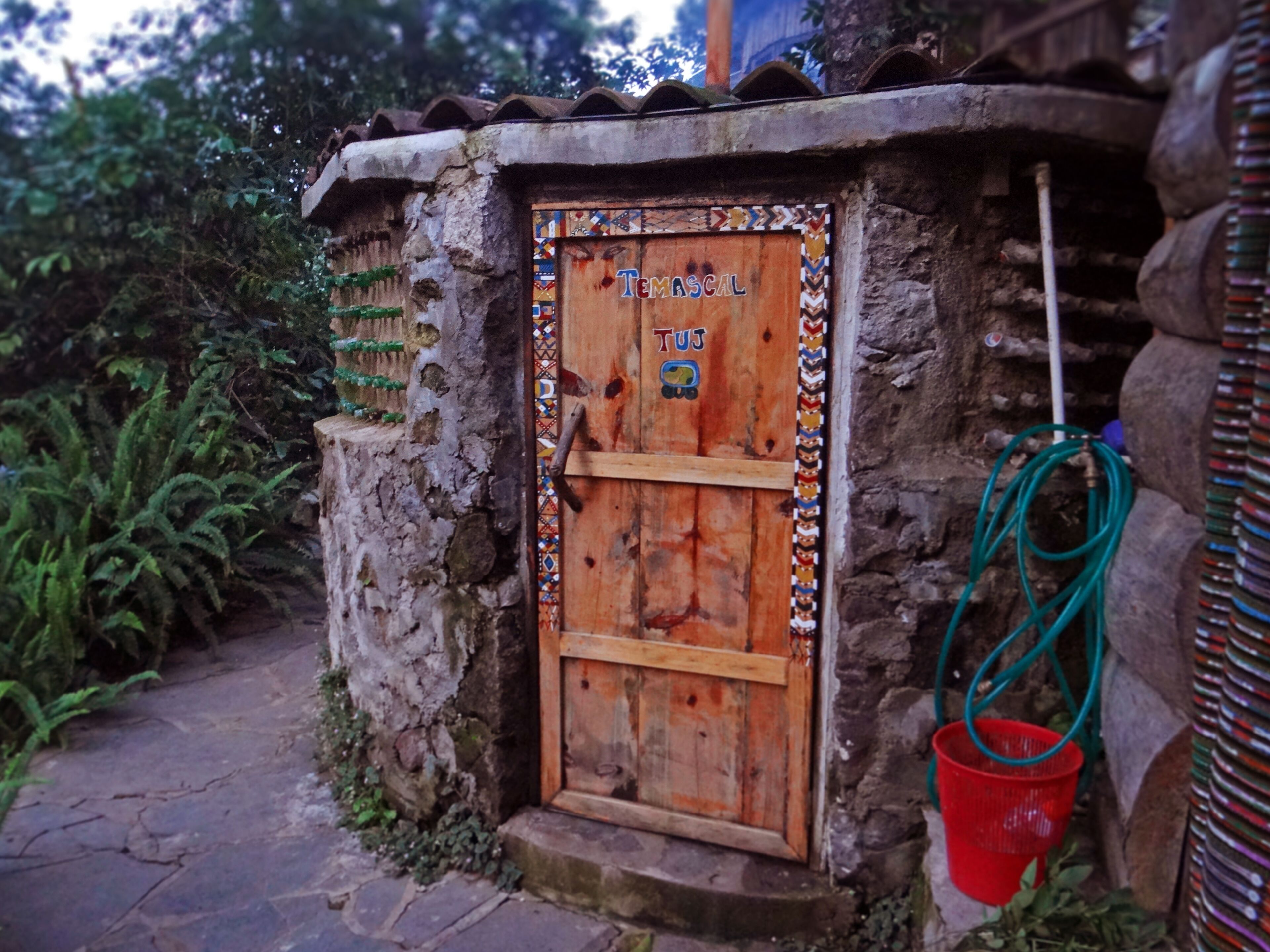 A rustic wooden door with a colorful border and a small window, set in a stone wall with a metal roof.