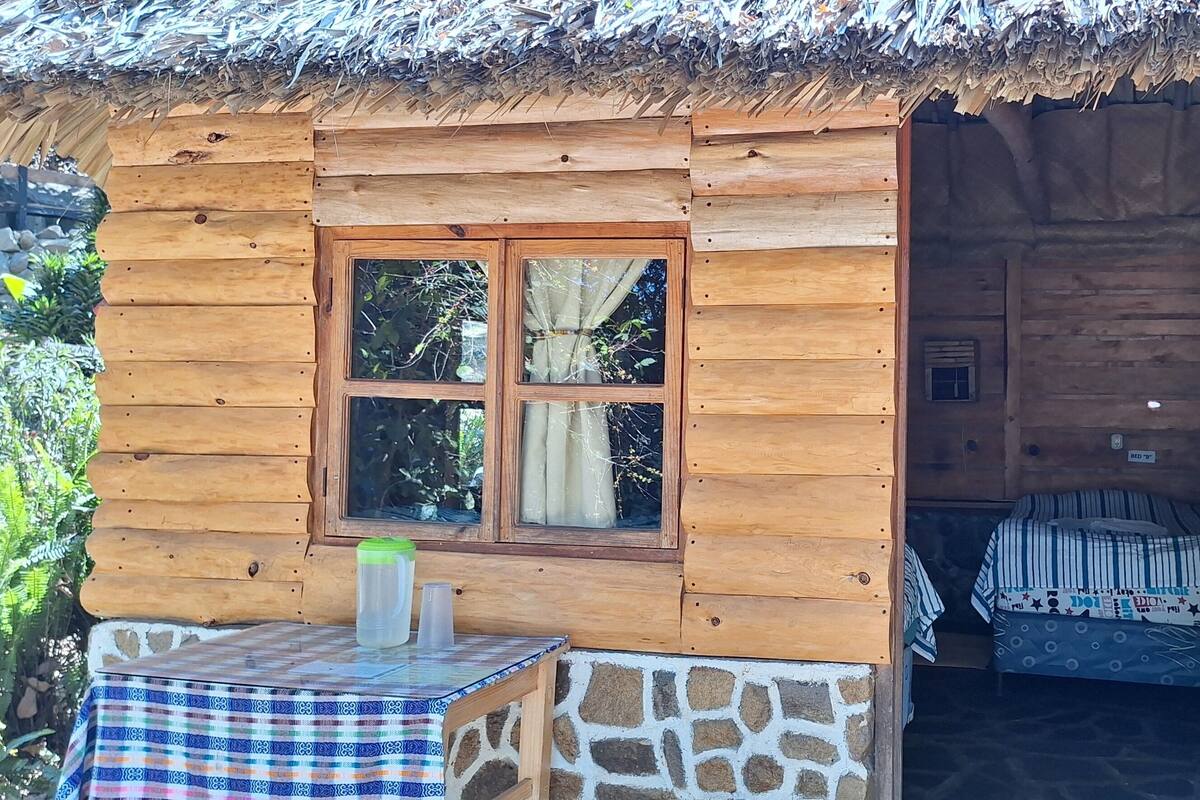 Een houten chalet met rieten dak, een raam met gordijnen, een bed met beddengoed met dessin, een tafel met geruit tafelkleed en een brandblusser aan de muur.