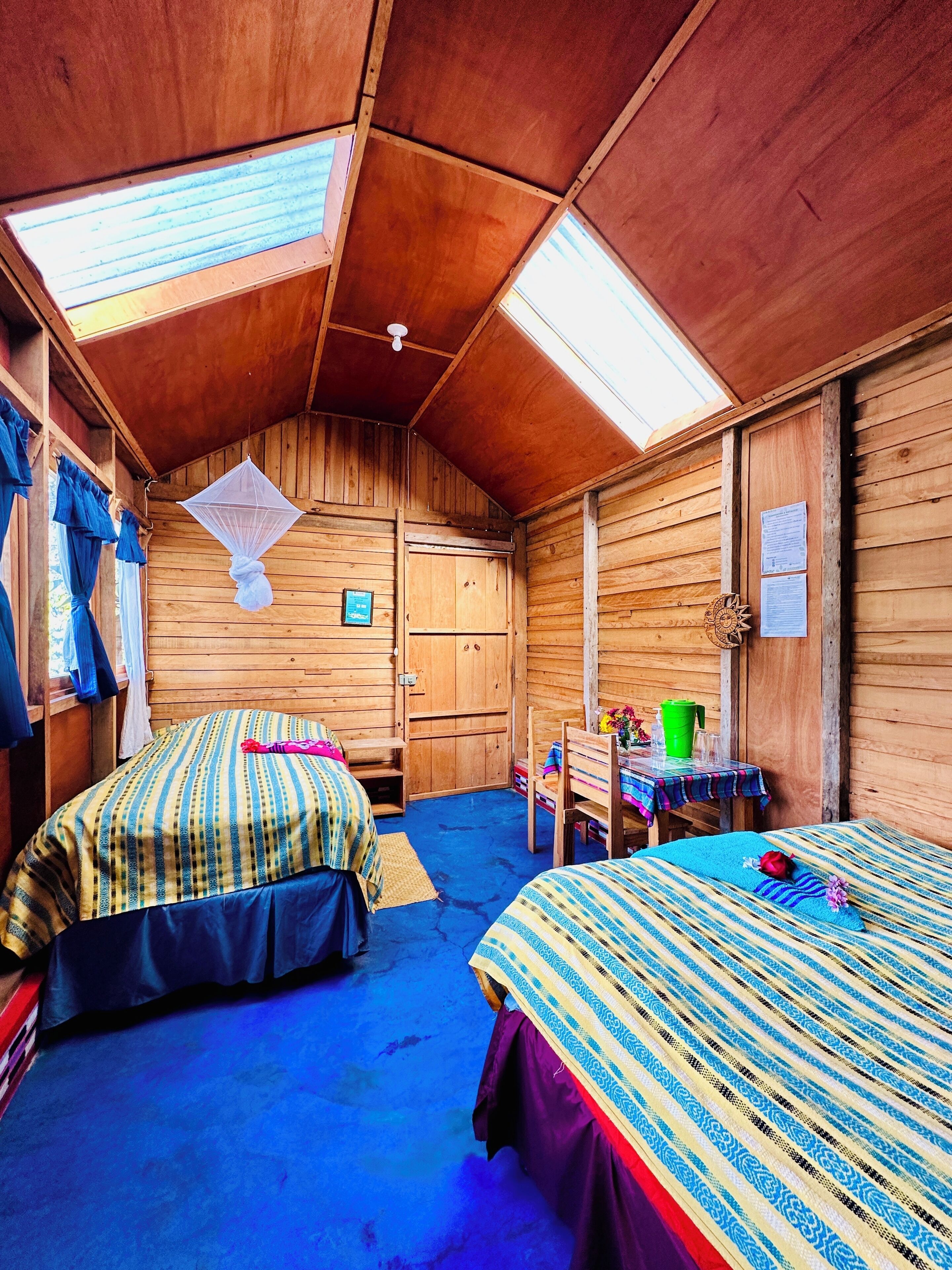 Een knus vakantiehuisje met twee bedden, een kleine tafel met een groene beker en glaasjes, en een houten plafond met dakramen.