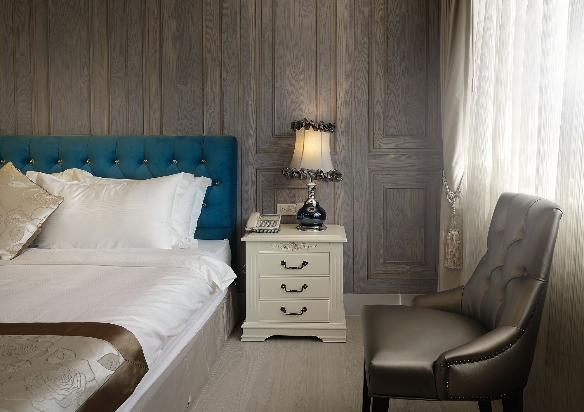 A neatly arranged hotel room with a blue upholstered headboard, a white bedside table with drawers, a leather armchair, and a wall-mounted telephone.