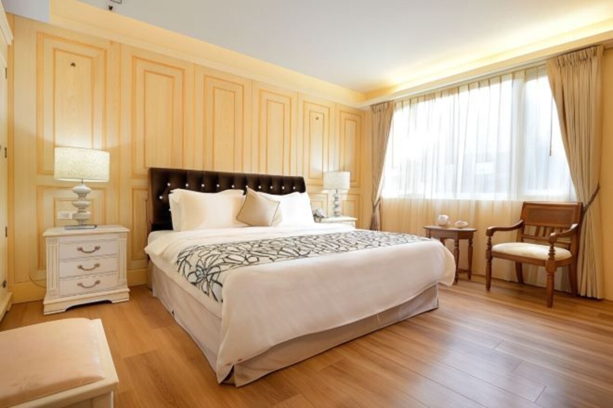 Ein Hotelzimmer mit einem großen Bett, Nachttischen, einem Sessel und einem Fenster mit Vorhängen.