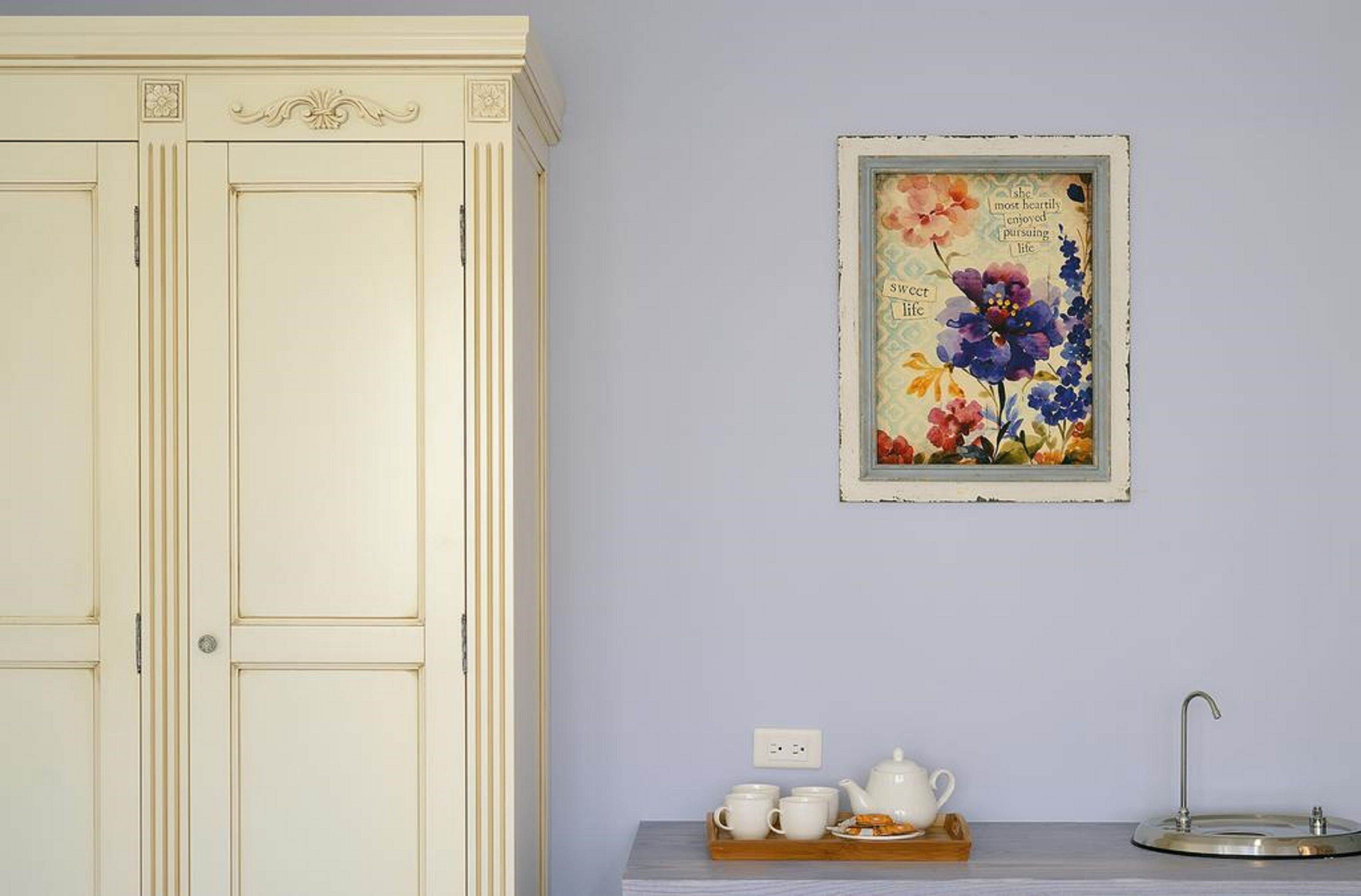 Una habitación con un armario color crema, una obra de arte floral enmarcadas en la pared, una mesita con una bandeja de tazas de té blancas y una tetera, y un lavabo.