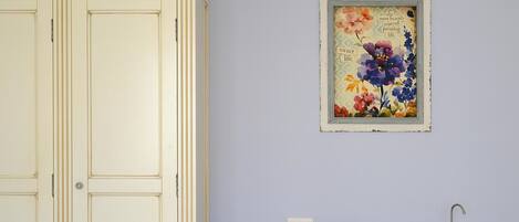 Una habitación con un armario color crema, una obra de arte floral enmarcadas en la pared, una mesita con una bandeja de tazas de té blancas y una tetera, y un lavabo.