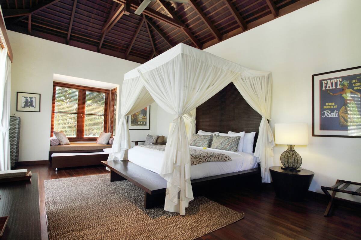 A bedroom with a four-poster bed, a bench, a bedside table, a lamp, and a framed picture on the wall.