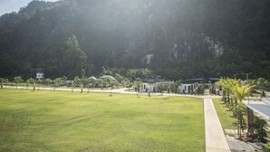 A large grassy field with a pathway leading to a building complex surrounded by a forested mountain. - Phuphayot Resort (Huai Yot)