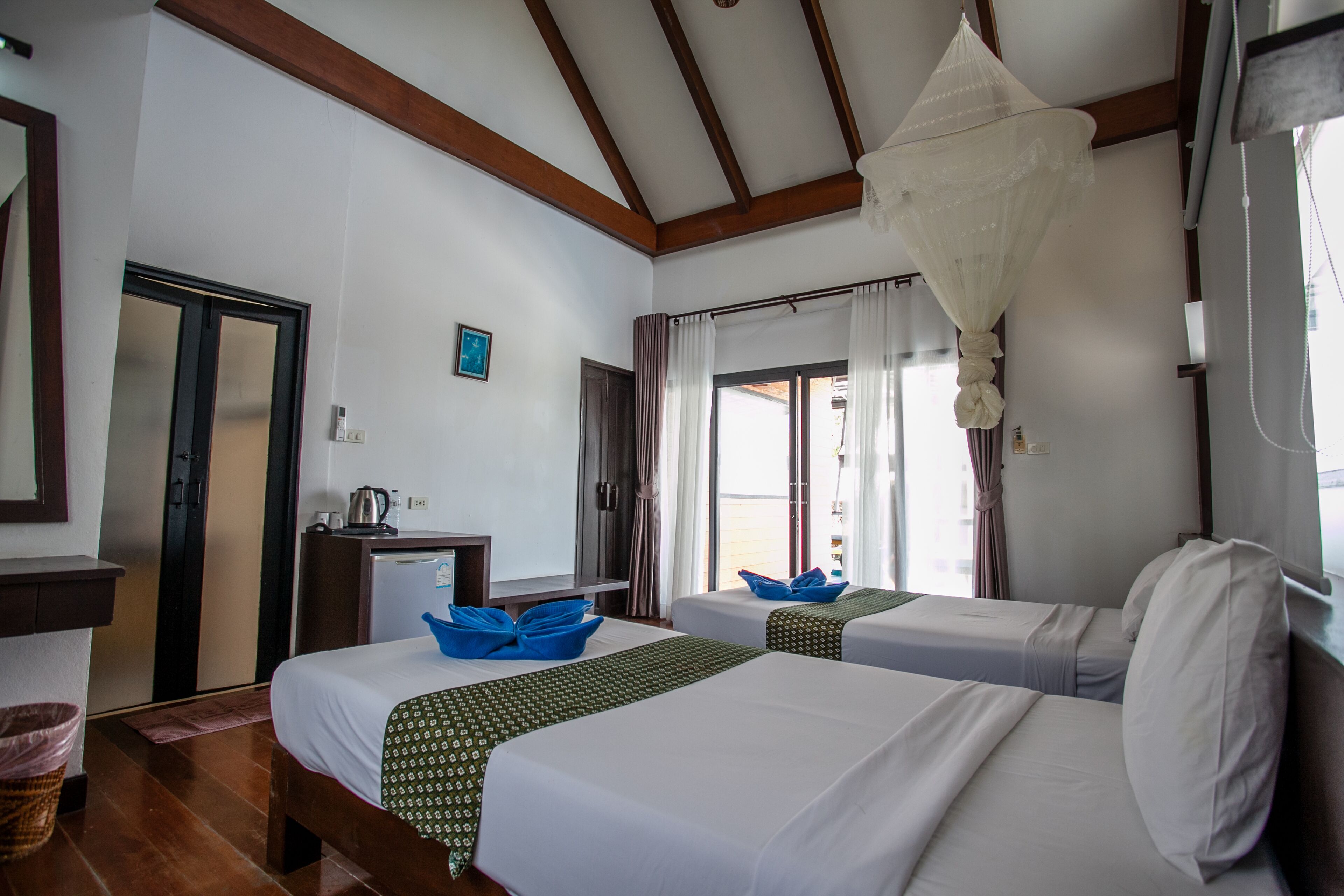A hotel room with two beds, a desk with a kettle, a hanging light fixture, and a view of the beach through the window.