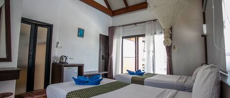A hotel room with two beds, a desk with a kettle, a hanging light fixture, and a view of the beach through the window.
