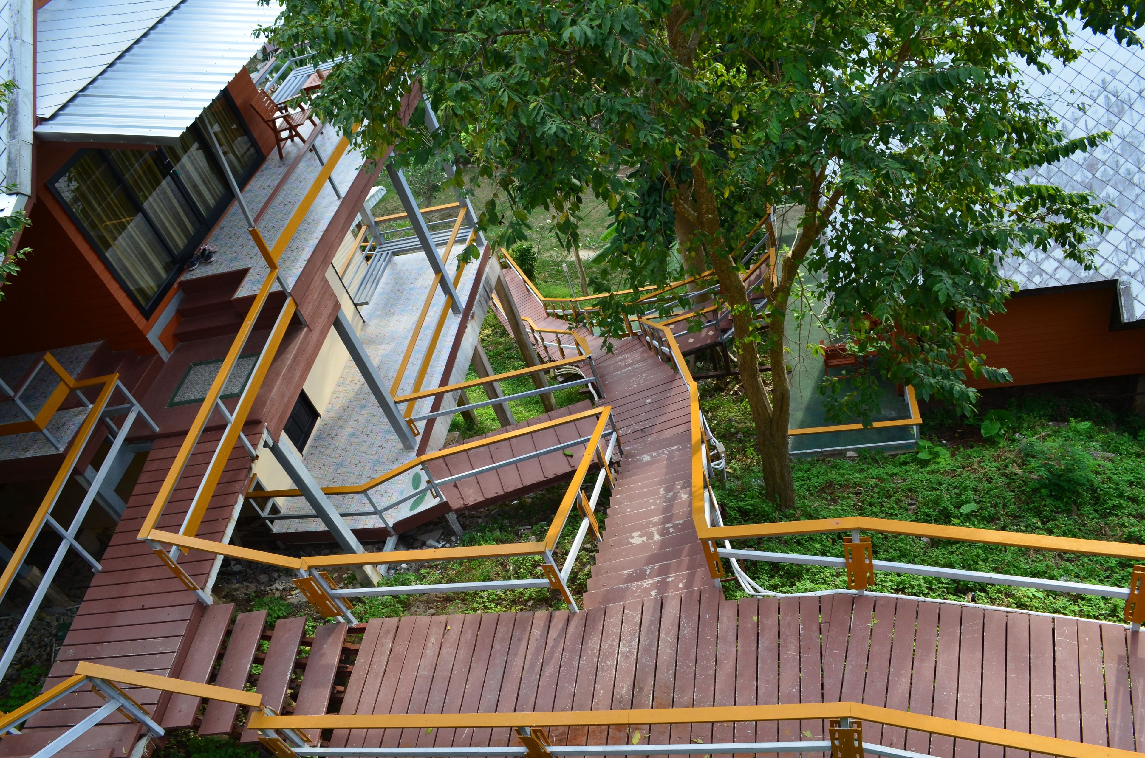 A multi-level wooden staircase with yellow handrails, surrounded by greenery and a building with a sloped roof.