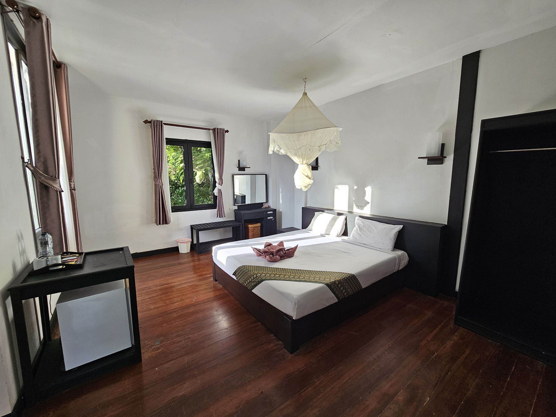 A hotel room with a large bed, bedside tables, a desk, a chair, a window with curtains, and a hanging light fixture.