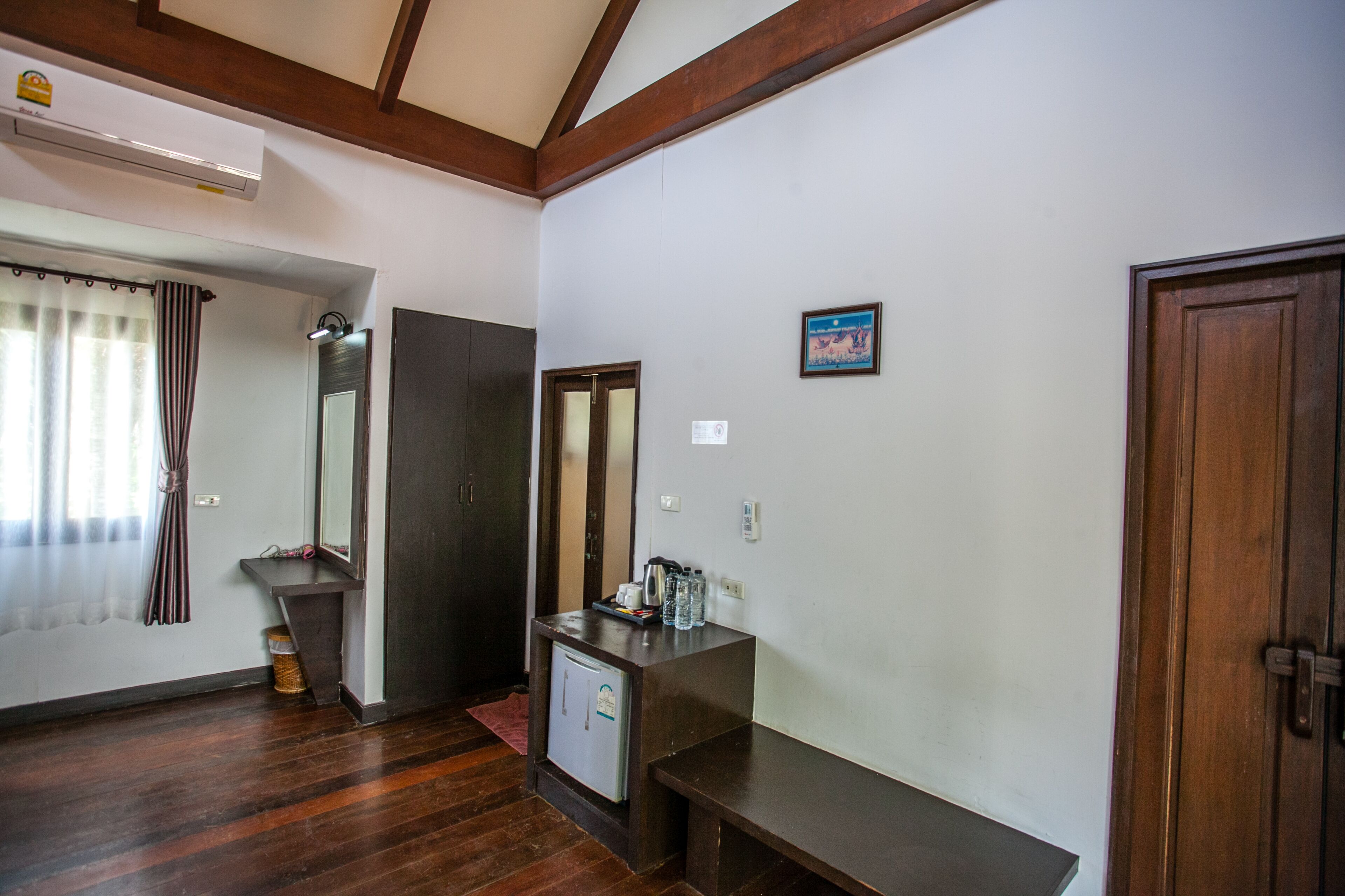 A hotel room with a wooden floor, a small desk with a mirror, a refrigerator, and a wooden door.