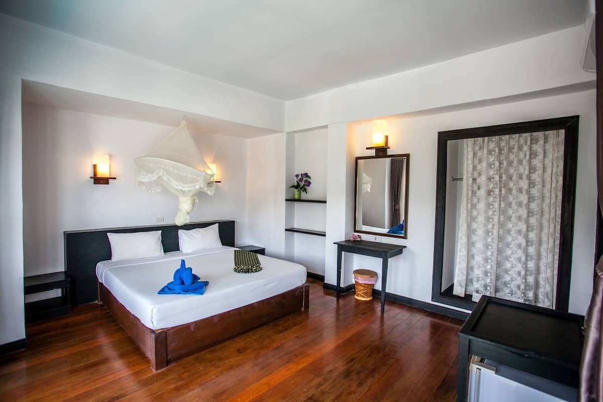 A hotel room with a bed, bedside tables, a nightstand, a mirror, a window with curtains, and a wall-mounted light fixture.