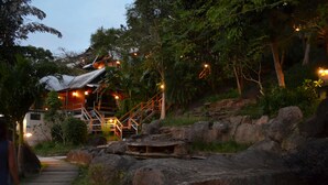 A rustic lodge with a wooden staircase leading to a thatched-roof building, surrounded by lush greenery and large rocks.