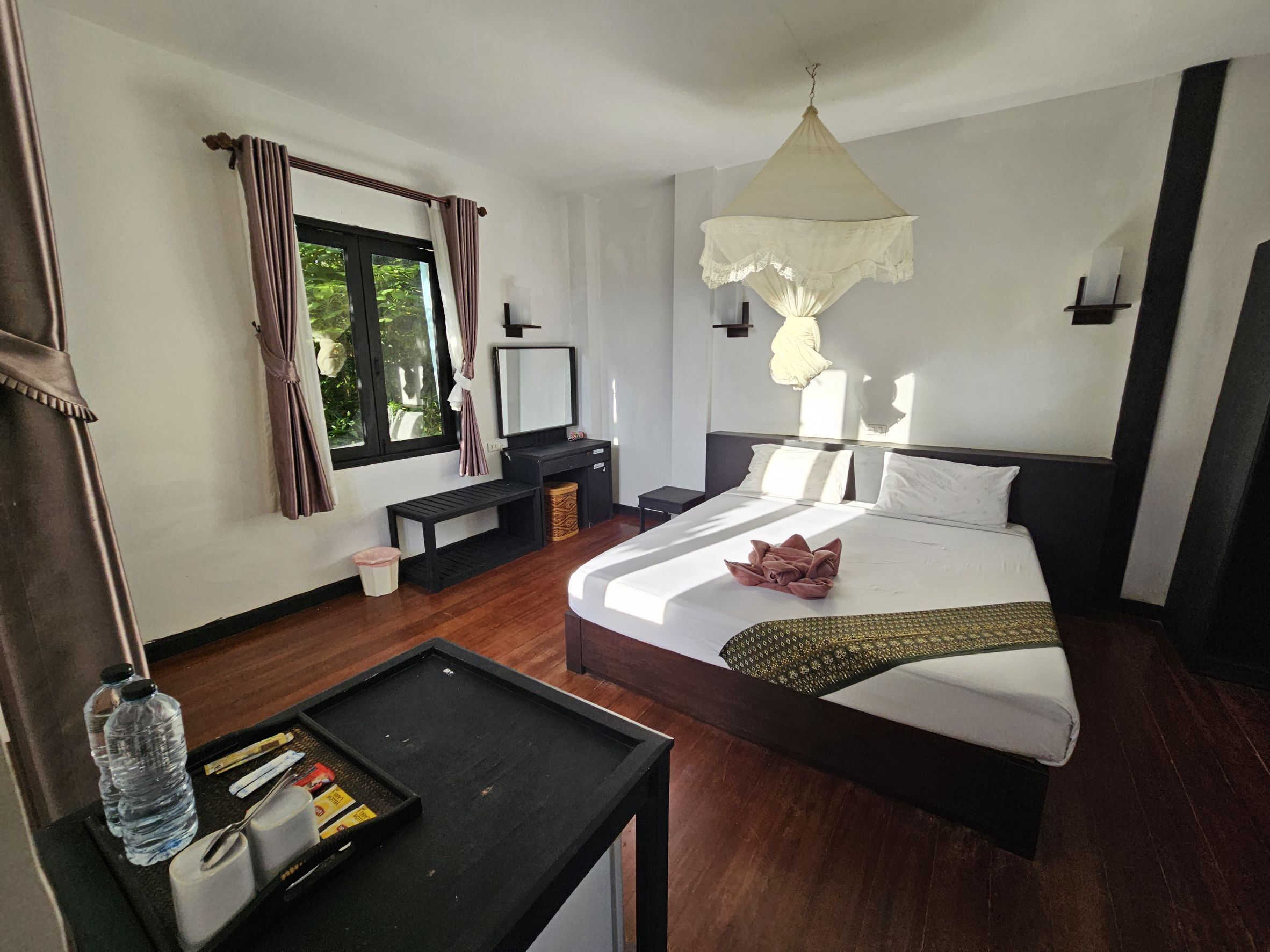 Una habitación de hotel con cama, mesita de noche, banco, ventana con cortinas y lámpara colgante.