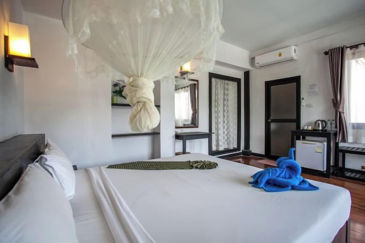 A hotel room with a bed, a bedside table, a lamp, an air conditioning unit, and a window with curtains.