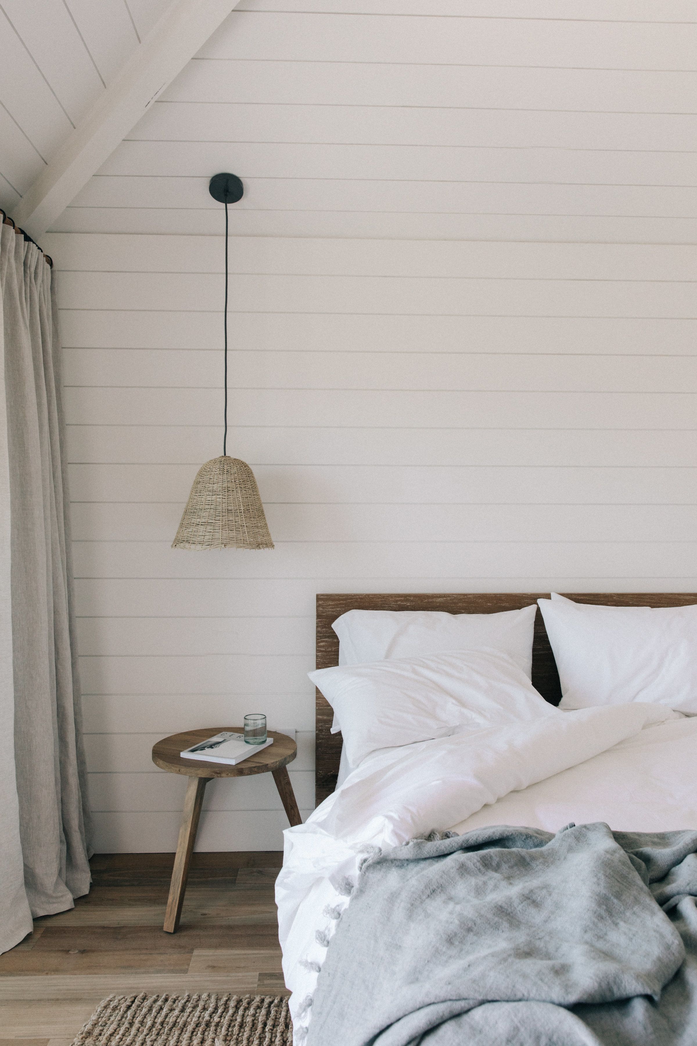 A bedroom with a wooden bed, white bedding, a woven pendant light, a small wooden side table with a glass, and a wooden floor.