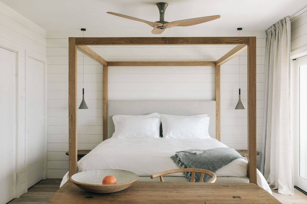 Ein Schlafzimmer mit einem hölzernen Baldachinbett, einem Holztisch mit einer Schüssel und Obst, sowie einem Deckenventilator.