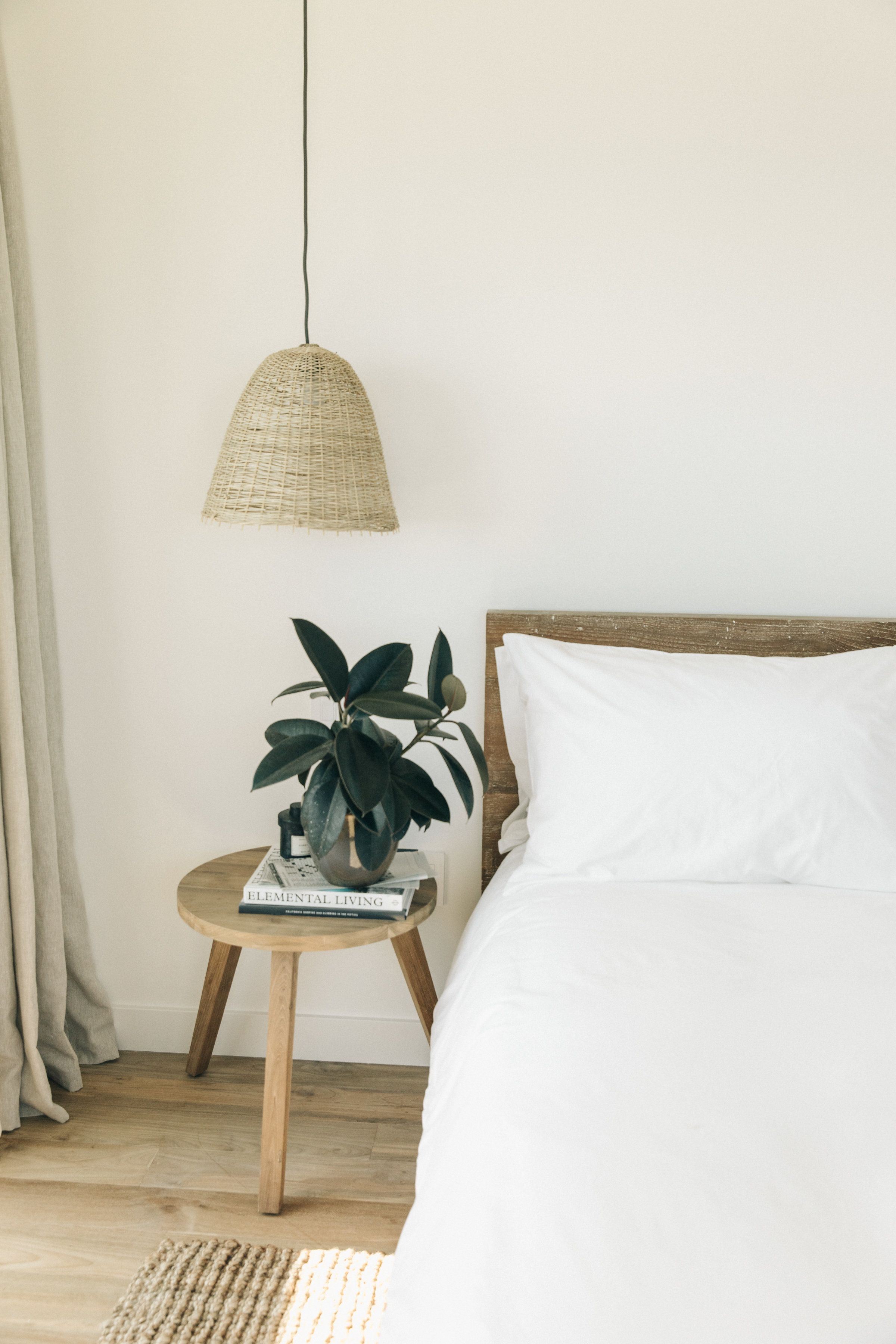 A bedroom with a bed, a bedside table with a plant, a book, and a woven pendant light.