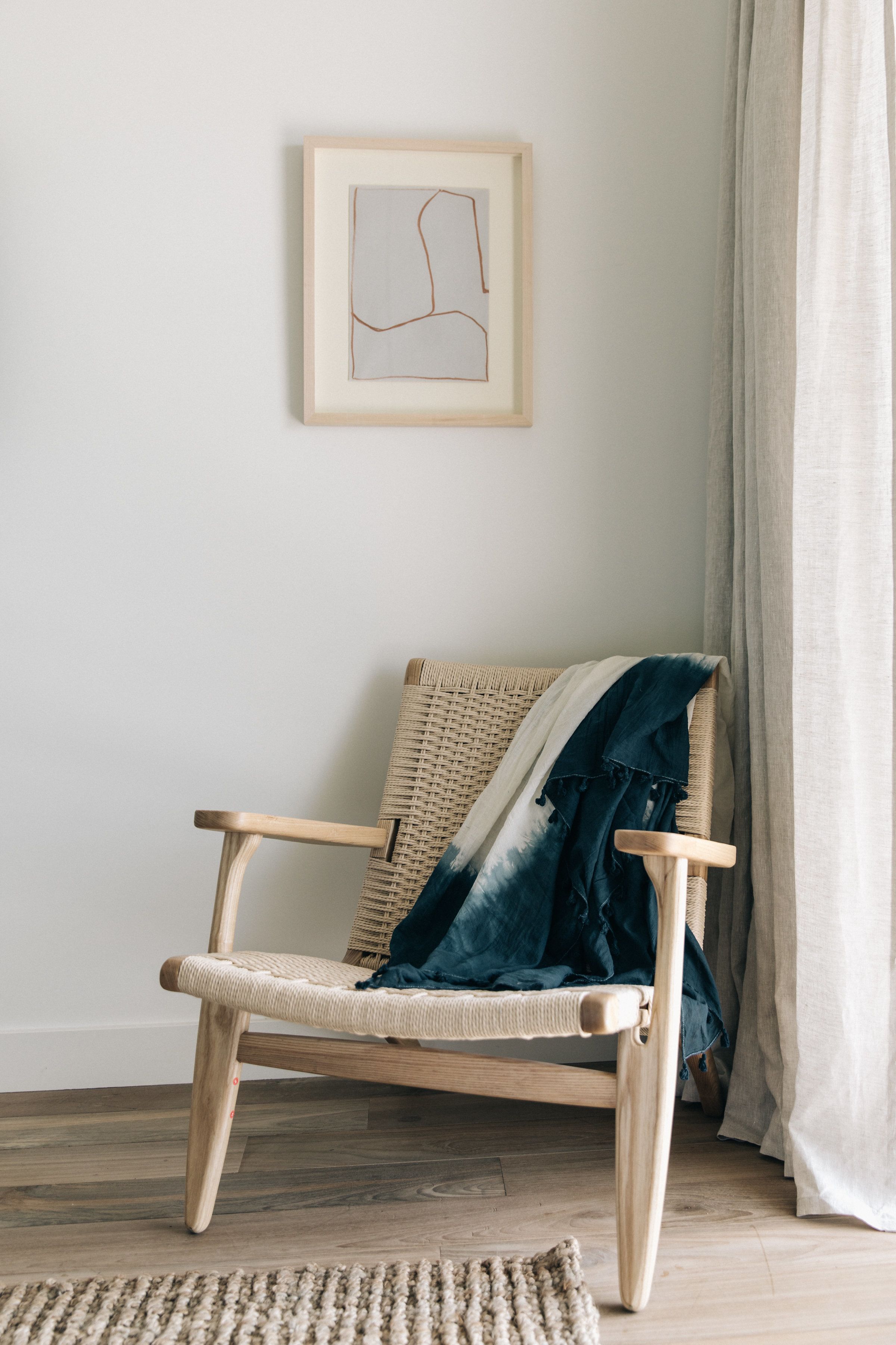 A wicker chair with a blue blanket, a wooden floor, and a framed abstract artwork on the wall.
