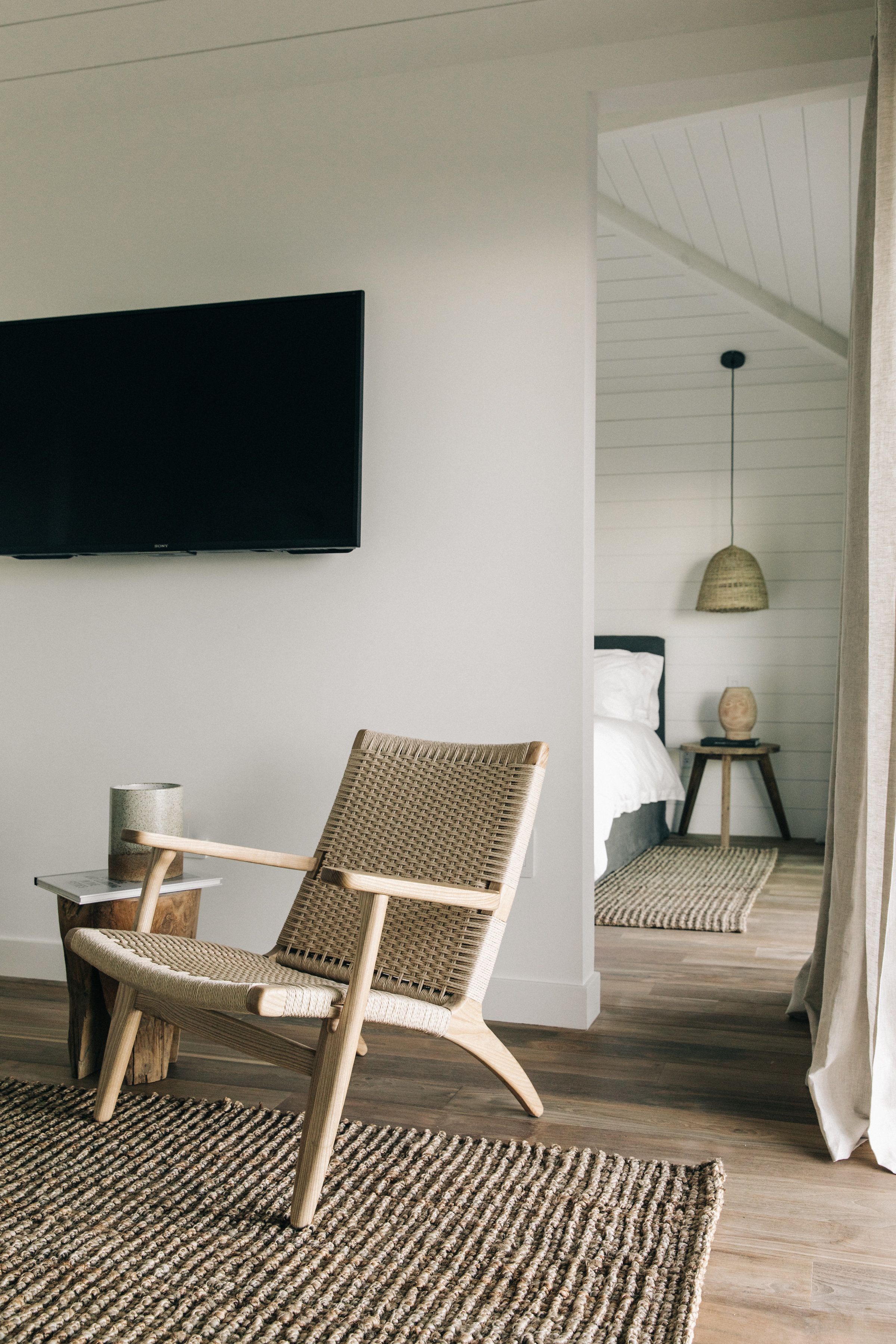 Habitación con un televisor de pantalla plana grande montado en la pared, una silla de mimbre, una mesita redonda con una vela, y una cama con cabecera blanca al fondo.