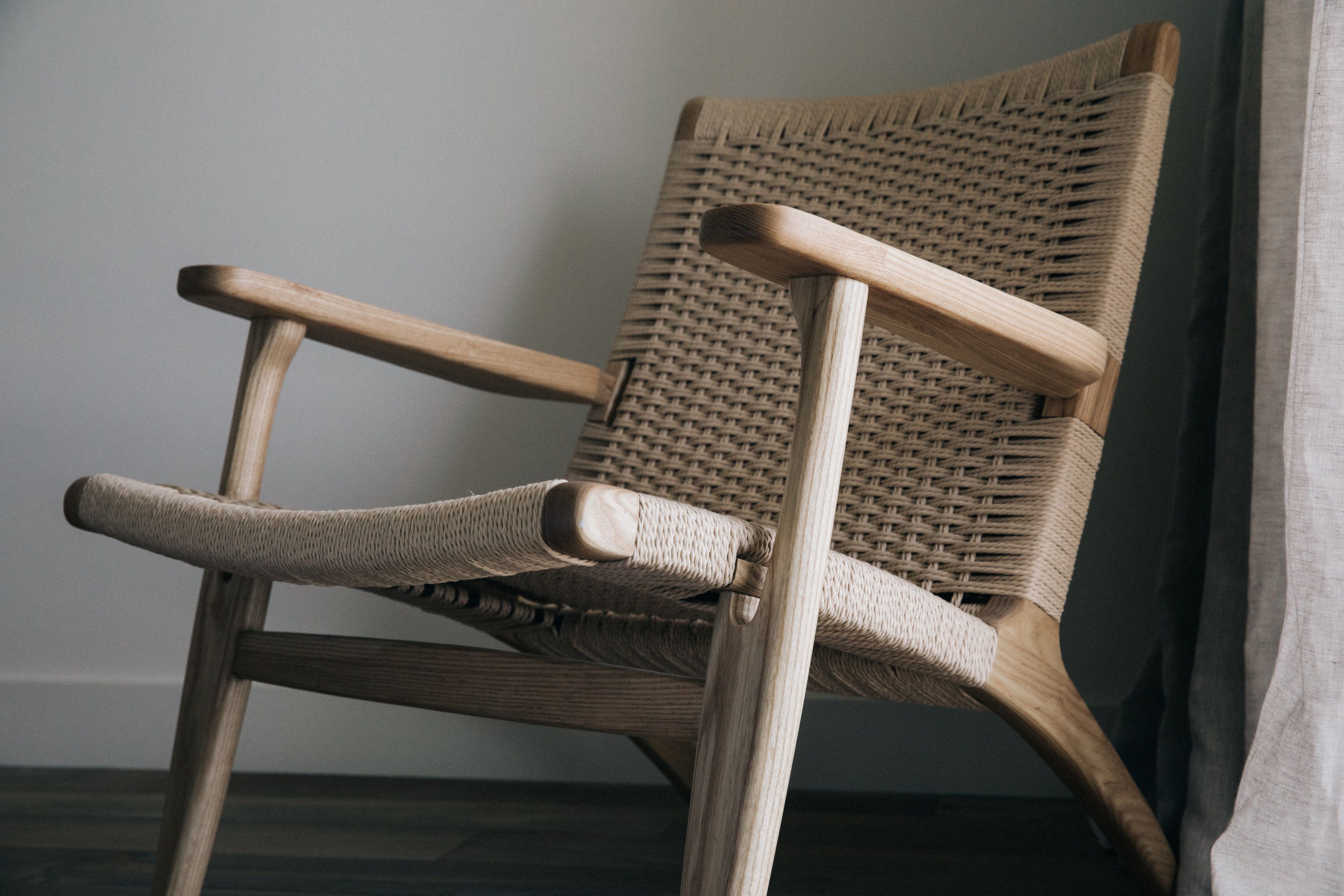 A wicker chair with wooden armrests and legs, positioned against a plain wall.