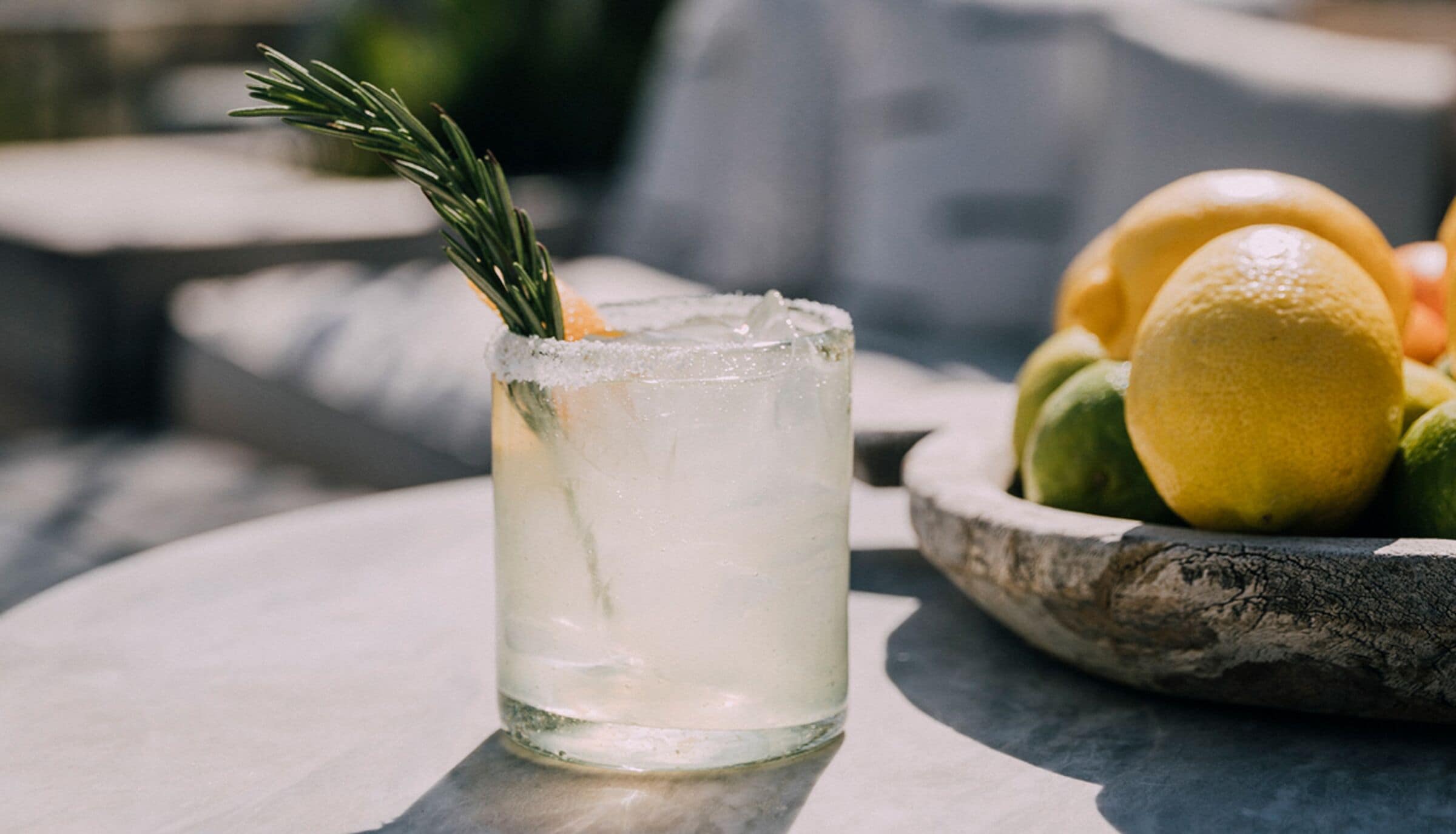 A clear glass with a garnish, placed on a table with a bowl of fruit in the background.
