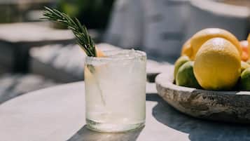 A clear glass with a garnish, placed on a table with a bowl of fruit in the background.