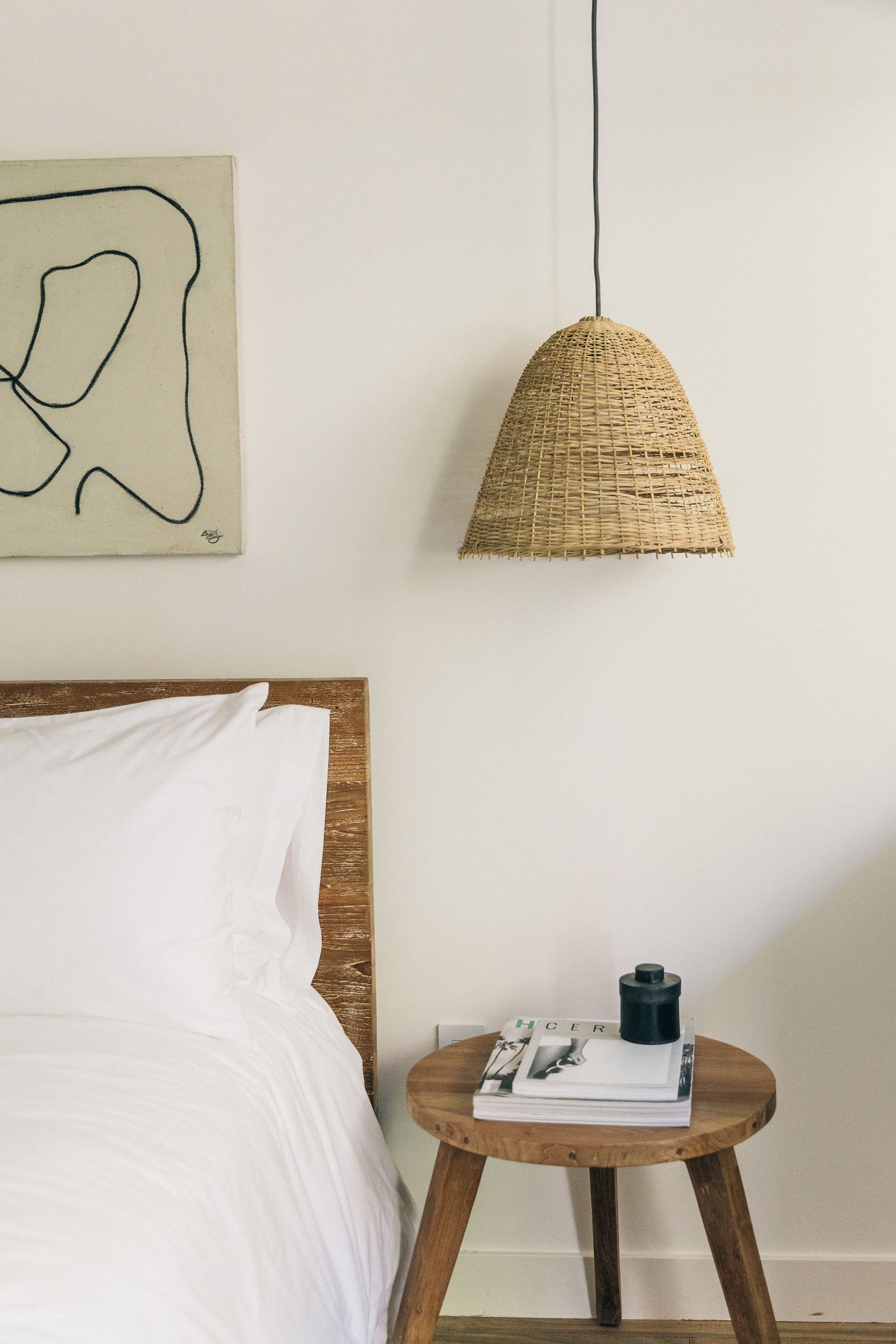 A bedroom with a wooden headboard, a white bedspread, a woven pendant light, and a small wooden table with a magazine and a black object on it.