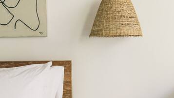 A bedroom with a wooden headboard, a white bedspread, a woven pendant light, and a small wooden table with a magazine and a black object on it.
