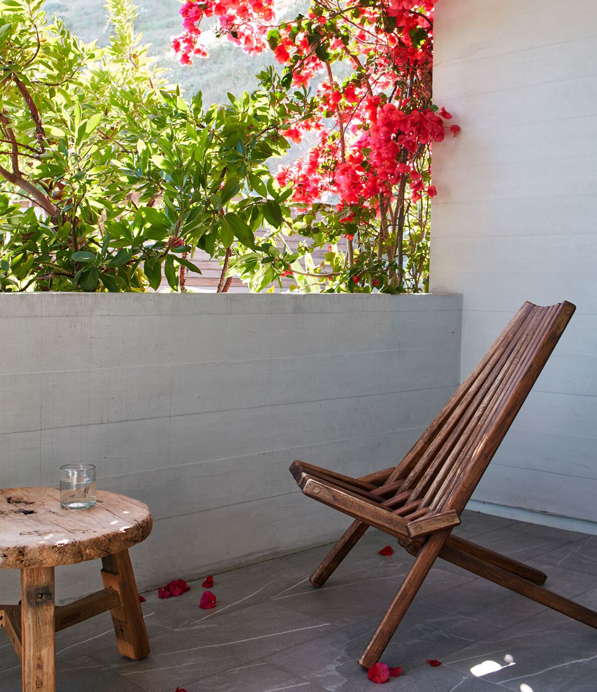 A wooden chair and a small round table with a glass of water on a balcony with a white wall and a plant with pink flowers.