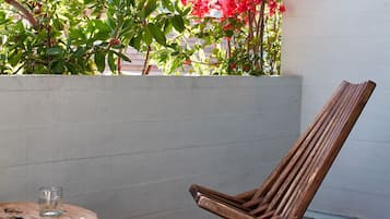 A wooden chair and a small round table with a glass of water on a balcony with a white wall and a plant with pink flowers.