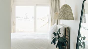 A bedroom with a large bed, a wooden floor, a round wooden table with a plant, and a hanging light fixture.