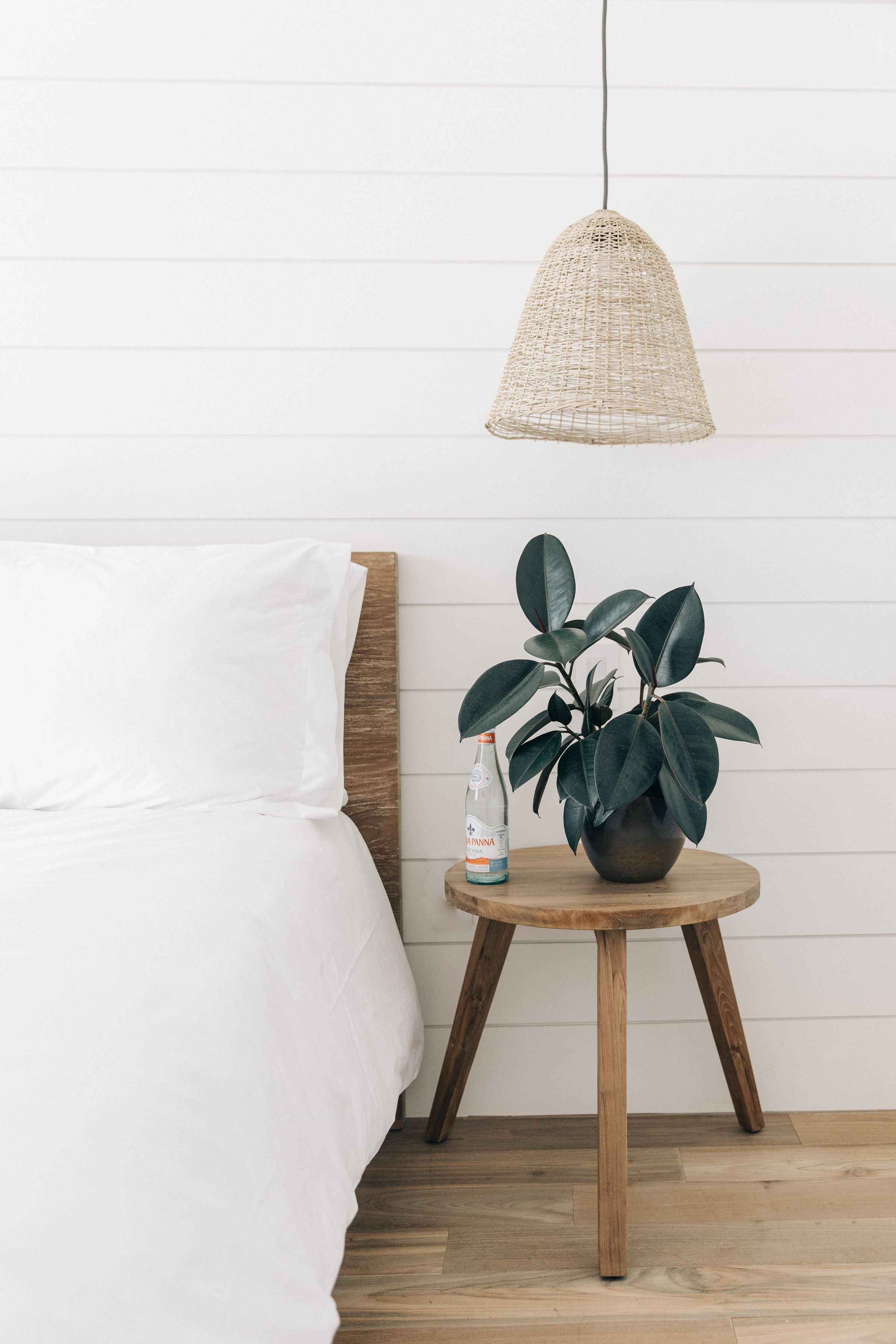 Uma cama com roupa de cama branca, uma mesa redonda de madeira com uma planta em vaso e uma garrafa, e uma luminária pendente trançada.