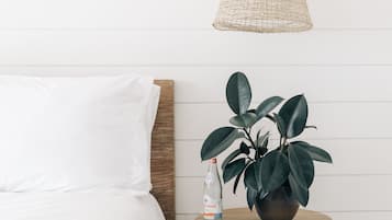 Uma cama com roupa de cama branca, uma mesa redonda de madeira com uma planta em vaso e uma garrafa, e uma luminária pendente trançada.