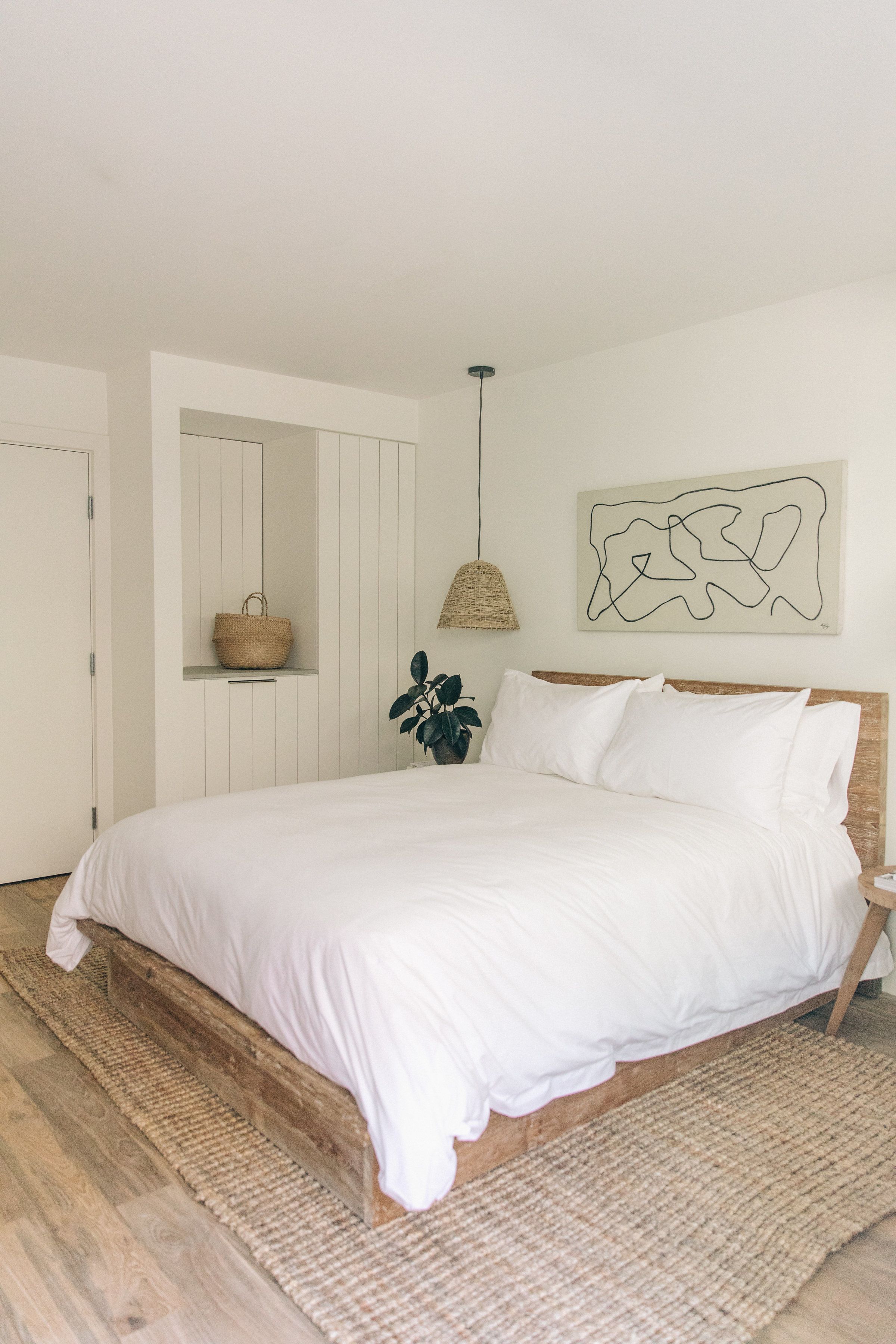 Un dormitorio con una cama grande, cabecera de madera, alfombra tejida, lámpara colgante, estante con cesto y una planta.
