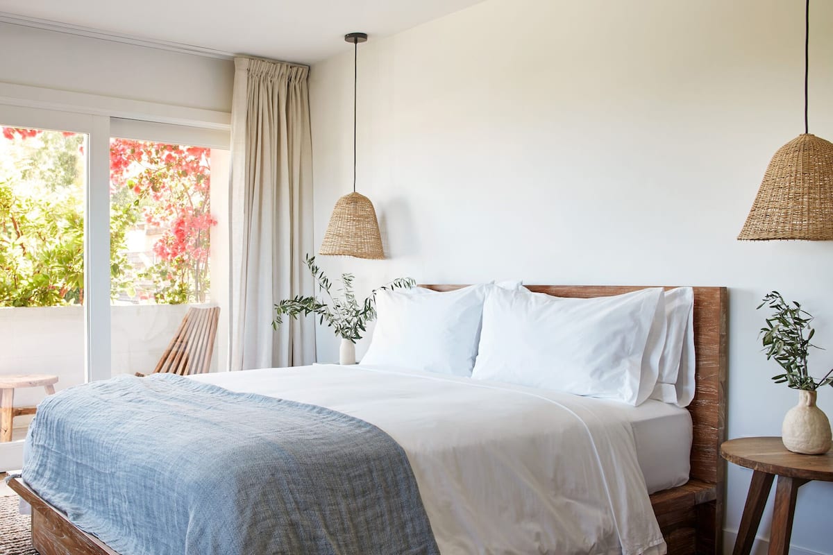 Ein Schlafzimmer mit einem großen Bett, einem hölzernen Kopfstück, einem Nachttisch, einer Pendelleuchte und Blick auf Grün.