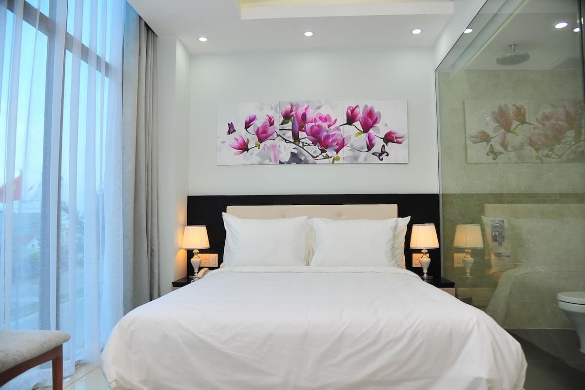 Une chambre d’hôtel avec un grand lit, deux tables de chevet avec des lampes, une grande fenêtre avec des rideaux légers et un tableau représentant des fleurs roses accroché au mur.