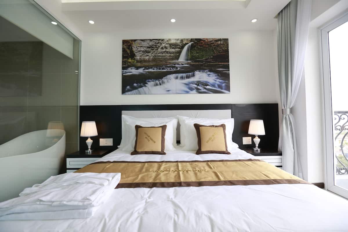 Une chambre d’hôtel avec un grand lit, deux tables de chevet avec des lampes, un tableau représentant une cascade accroché au mur, et une vue sur l’extérieur par une fenêtre.