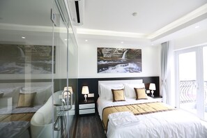 A modern hotel room with a large bed, a glass partition, a nightstand, and a view of a waterfall through the window. - Golden Dragon Hotel (Gia Tran)