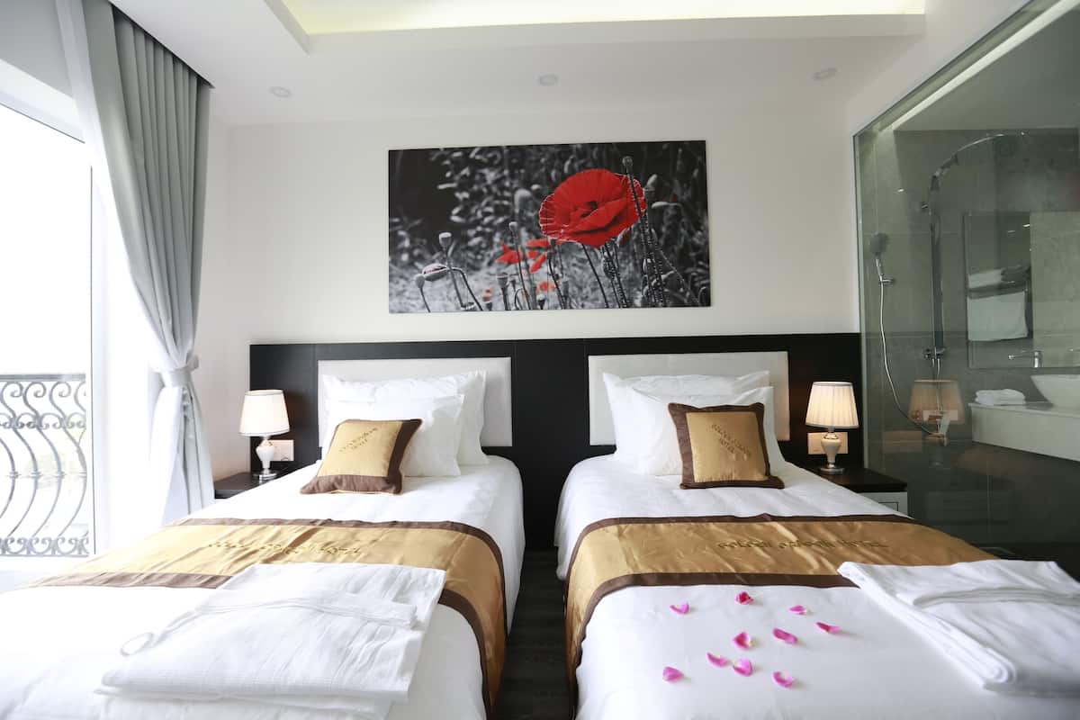 Une chambre d’hôtel avec deux lits, une grande fenêtre avec des rideaux, un tableau représentant des fleurs rouges, une douche en verre et une salle de bain avec un lavabo et un miroir.