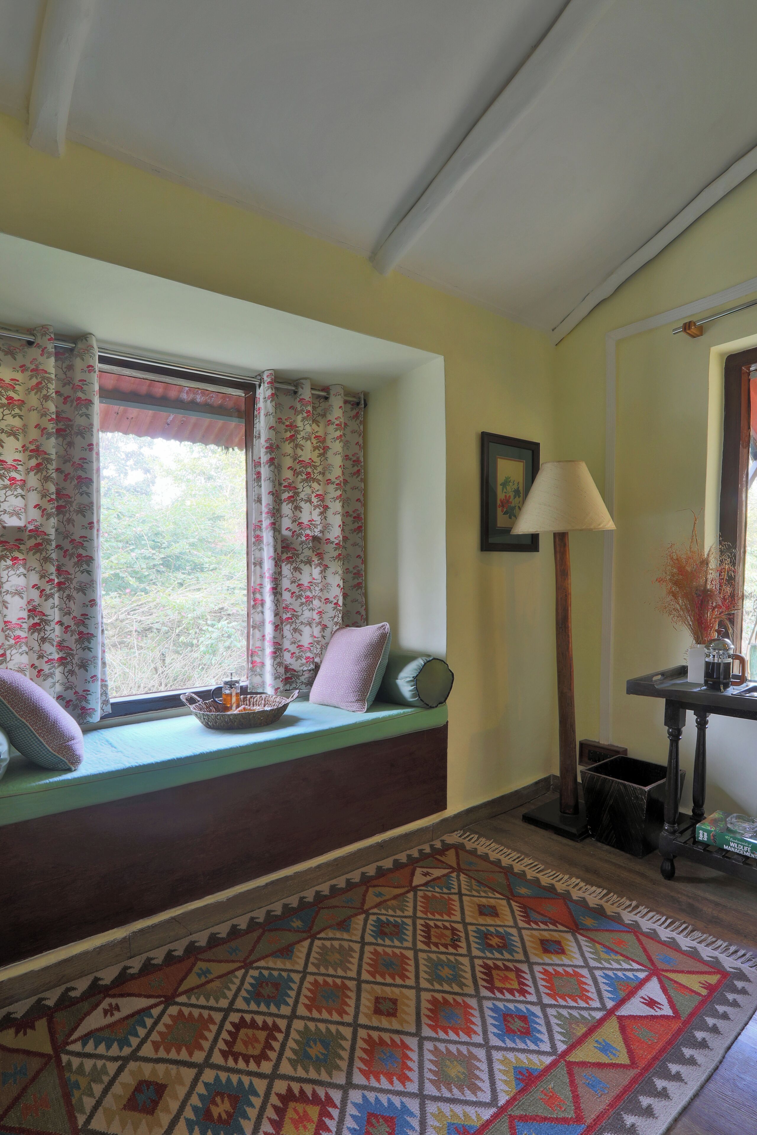 Una habitación con un asiento junto a la ventana, un tapete estampado, una lámpara y un cuadro en la pared.