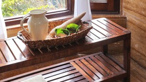 A wooden table with a woven basket containing a pitcher and a green plant, a white towel, and a window with a view of greenery. - Jim's Jungle Retreat (Ramnagar)