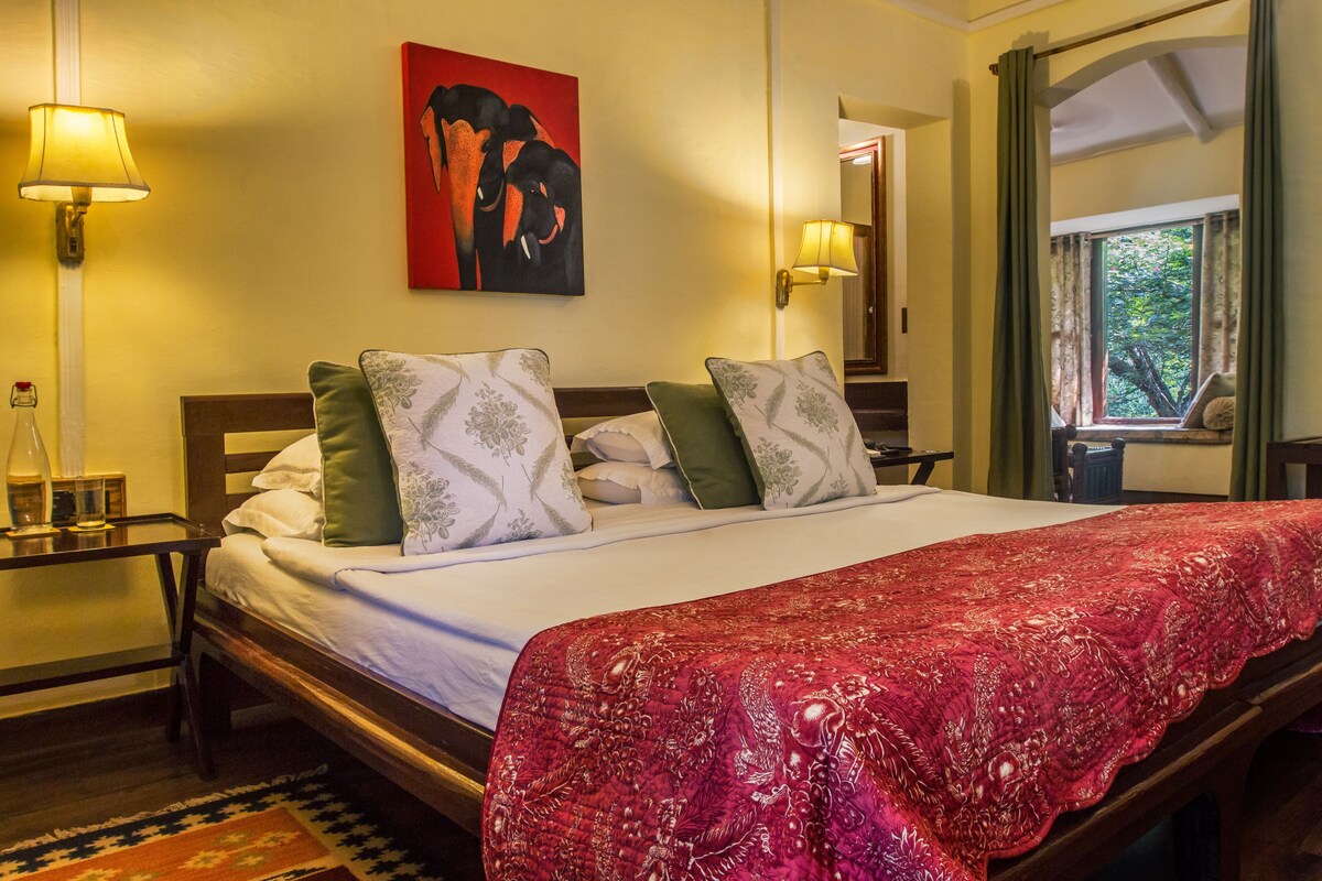 Una cama bien hecha con una colcha roja, dos mesitas de noche con lámparas, un cuadro de elefantes y una ventana con cortinas.