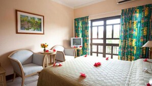 A hotel room with a bed, two chairs, a small table with a vase of flowers, a television, and a window with curtains. - Gregoire's Apartments (La Digue)