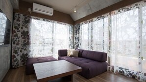 A modern living room with a sofa, a wooden coffee table, and large windows with floral curtains. - Condominio Makishi Annesso (Naha)