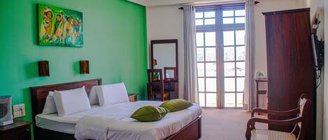 A bedroom with a green wall, a large bed with white and green pillows, a wooden chair, a wooden wardrobe, and a window with curtains.