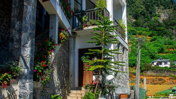 Une maison à deux étages avec une façade en pierre, un balcon en bois et un jardin luxuriant où poussent différentes plantes et fleurs.