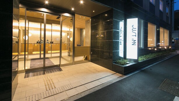 A modern hotel entrance with glass doors, a visible reception area, and a sign reading "JUST IN." - Just Inn Premium Nagoya Station (Nagoya)
