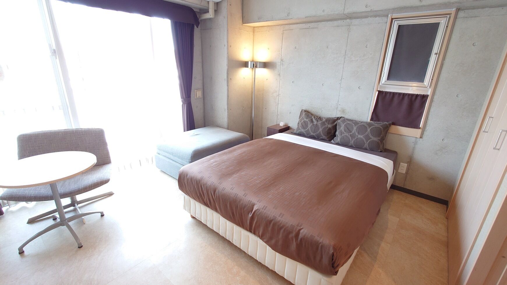 A hotel room with a bed, a chair, a small table, and a window.