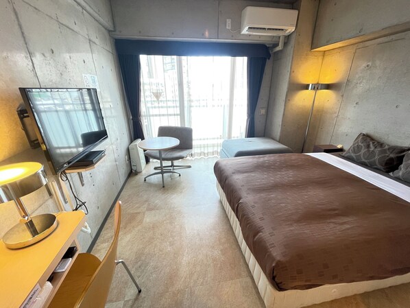 A hotel room with a bed, a desk with a lamp, a chair, a TV, and a window with curtains. - Condominio Makishi (Naha)