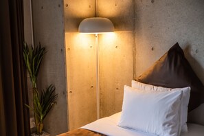 Una habitación de hotel con cama, lámpara de pie, planta en maceta y almohada.